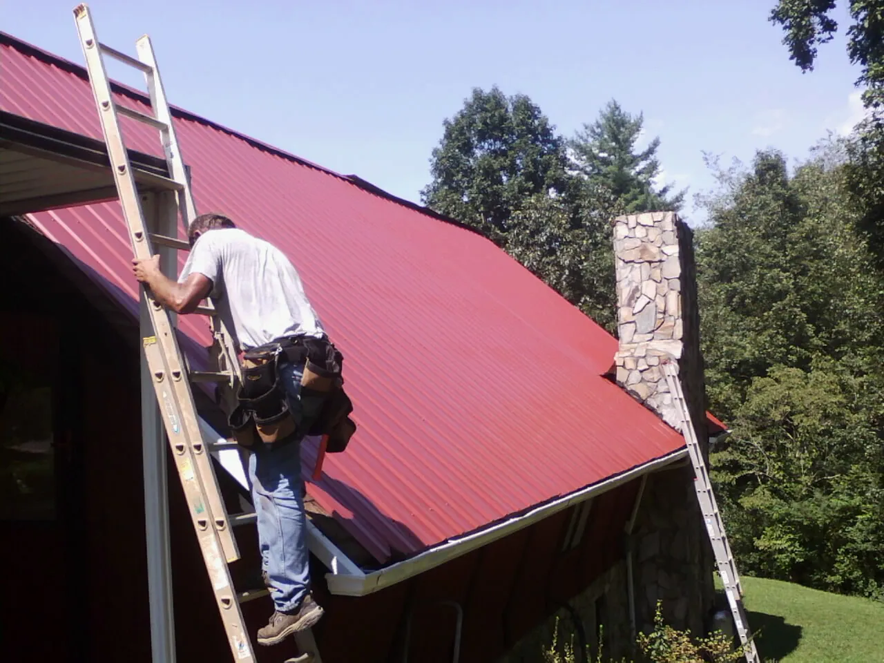 Roofer on a ladder beside a red metal roof and stone chimney — Soffit & Fascia Repair by Roofing Brothers in Green Mountain Falls
